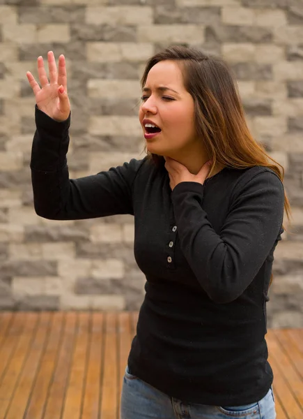 Joven hermosa mujer sintiéndose mal y agarrando su cuello con una mano y pidiendo ayuda con su otra mano, concepto de reanimación cardiopulmonar, en un fondo borroso — Foto de Stock