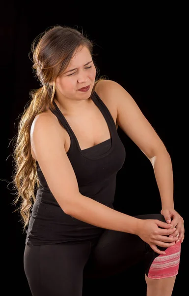 Beautiful young attractive sport woman holding injured knee suffering pain, in black background — Stock Photo, Image