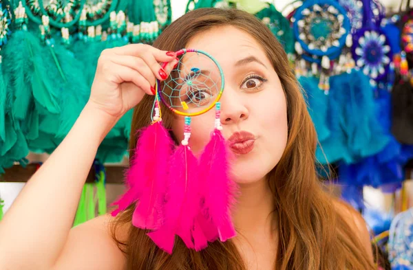 Close up of a beautiful smiling young woman pointing in from of her face a pink catchdreamers, in colorful market fabrics background — Stock Photo, Image