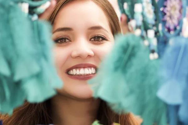 Primer plano de una mujer sonriente escondida detrás de un atrapasueños magenta, en un colorido fondo de mercado — Foto de Stock