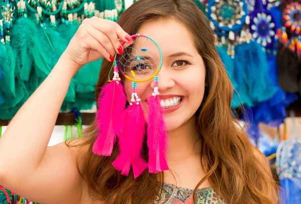 Acercamiento de una hermosa joven sonriente que señala desde su cara a un atrapasueños rosados, en telas coloridas de mercado de fondo — Foto de Stock