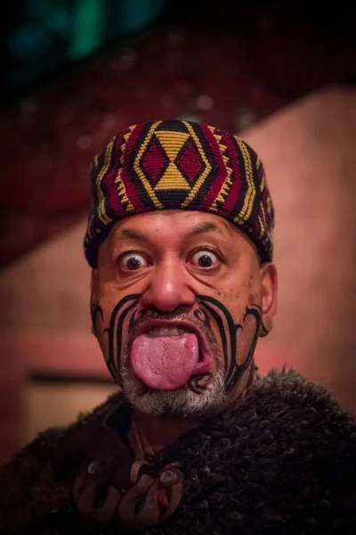 НОРТ-ОСТРОВ, НЬЮ-ЙОРК, 17 января 2017 года: мужчина маори, торчащий из языка, с традиционно татуированным лицом в традиционной одежде в Культурной деревне маори в шляпе маори — стоковое фото