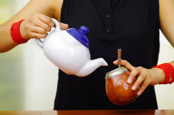 Woman holding white small jar pouring hot water into traditional cup with typical metal straw sticking up, preparing popular drink called mate — Stock Photo, Image