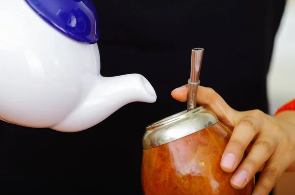 Femme tenant petit pot blanc versant de l'eau chaude dans une tasse traditionnelle avec de la paille métallique typique collant vers le haut, préparant une boisson populaire appelée mate — Photo