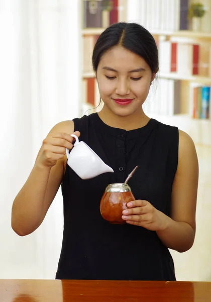 Mujer sosteniendo un pequeño frasco blanco vertiendo agua caliente en la taza tradicional con paja metálica típica que sobresale, preparando una bebida popular llamada mate. — Foto de Stock