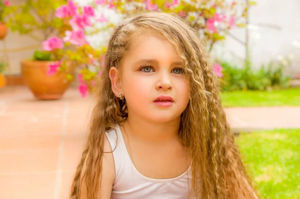 Portrait of a beautiful little girl preschool sitting on ground posing for camera, in a garden background — Stock Photo, Image