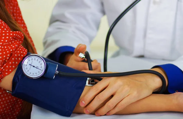 Primer plano de un médico joven usando el estetoscopio para escuchar el pulso mientras una mujer está con un tensiómetro en el brazo, en un fondo de la sala de consulta médica — Foto de Stock