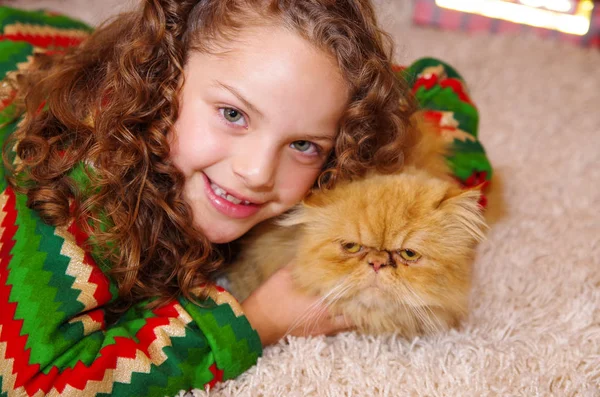 Krásná usměvavá drobná dívka, která nosí vánoční oblečení, objal její žlutá kočka na bílý koberec — Stock fotografie