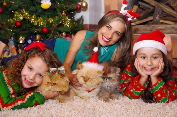 Belle famille portant des vêtements de Noël, embrassant ses chats, la fille bouclée avec une cravate rouge dans les cheveux pendant que la fille brune porte un chapeau de Noël à côté de sa mère, allongée sur un tapis blanc — Photo