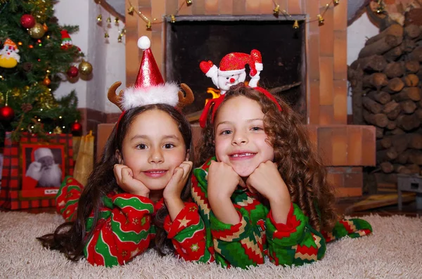 Twee mooie lachende litle meisjes dragen een kerst kleding tot op een wit tapijt, met een overdekt chimmey achtergrond — Stockfoto