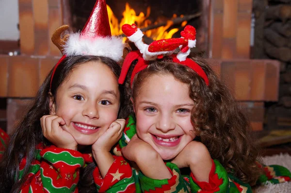 Close-up van twee mooie lachende litle meisjes dragen een kerst kleding tot op een wit tapijt, met een overdekt chimmey achtergrond — Stockfoto