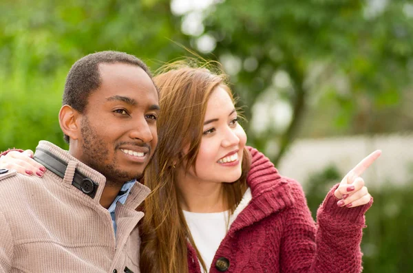 Hermosa y sonriente pareja joven interracial feliz en el parque, mujer señalando en algún lugar en un fondo del parque — Foto de Stock