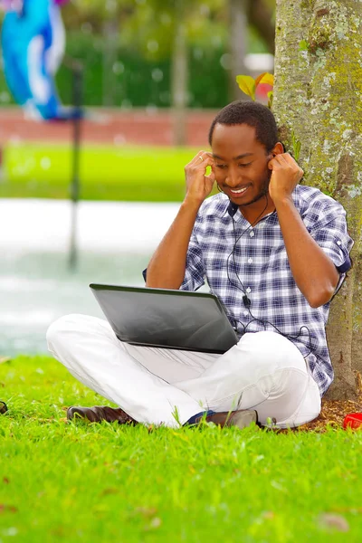 Junger schwarzer Mann sitzt auf grünem Gras und arbeitet an seinem Computer und hört Musik mit seinen Kopfhörern posiert seinen Rücken in einem Baum in der Stadt Quito ecuador — Stockfoto