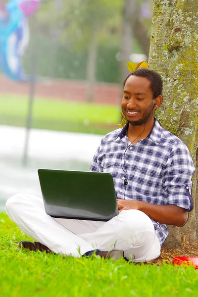 Joven hombre negro sentado sobre hierba verde y trabajando en su computadora posando su espalda en un árbol en la ciudad de Quito Ecuador —  Fotos de Stock