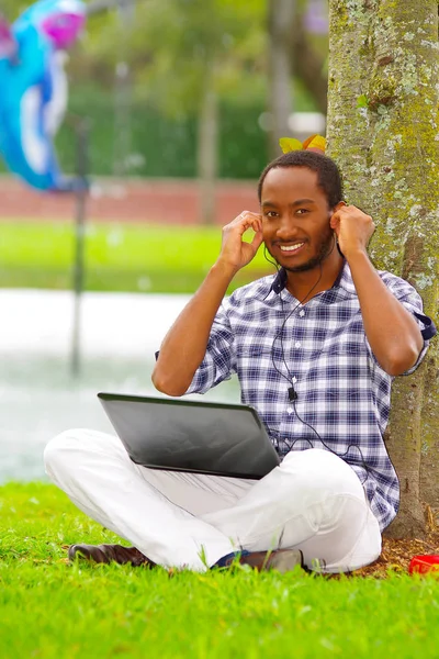 Jonge zwarte man op gras zitten en werken in zijn computer en muziek luisteren met zijn hoofdtelefoon poseren zijn rug in een boom in de stad van Quito-Ecuador — Stockfoto