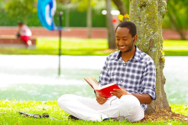 Junger schwarzer Mann sitzt auf grünem Gras, liest ein Buch und posiert mit dem Rücken in einem Baum in der Stadt Quito ecuador — Stockfoto