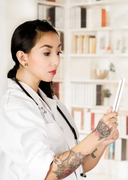 Beautiful tattooed young doctor holding in her hands a tablet reading a medical results from patients, in office background — Stock Photo, Image