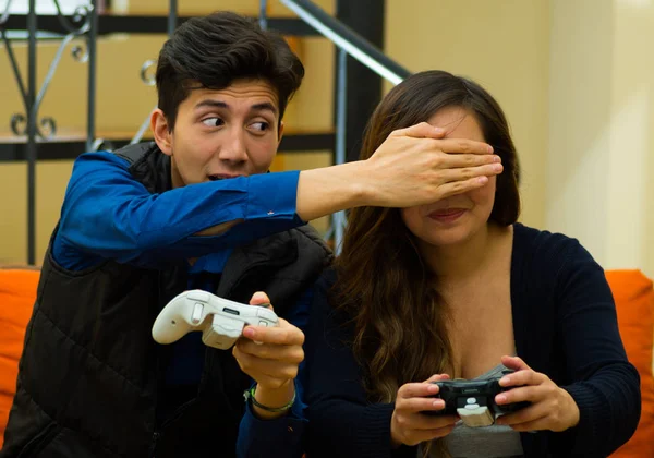 Jovem bonito jogando videogames no sofá, cobrindo os olhos de sua amiga, conceito sobre entretenimento em casa, jogos de vídeo — Fotografia de Stock