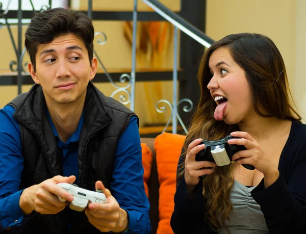 Красивый улыбающийся молодой человек играет в видеоигры с выражением победы на лице, в то время как его девушка проигрывает и торчит щипцы своему парню на диване, концепция о доме — стоковое фото