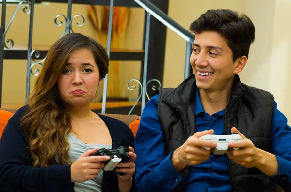 Красивый улыбающийся молодой человек играет в видеоигры с выражением победы на лице, пока его девушка проигрывает на диване, концепция домашнего развлечения, видеоигры — стоковое фото