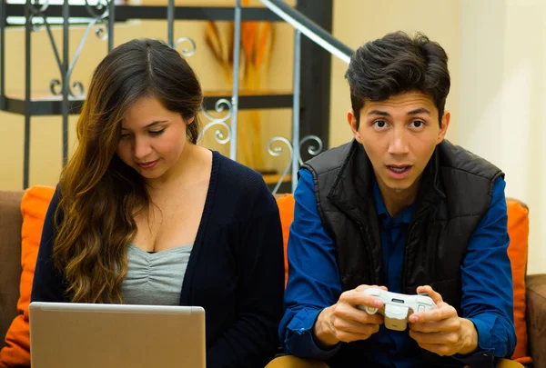 英俊的年轻男子玩电子游戏，而他的女朋友在她的电脑，在沙发上，工作家庭娱乐，视频游戏的概念 — 图库照片