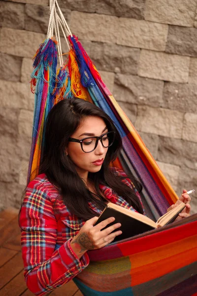 Vackra unga hipster kvinnan avslappnande och liggande i en färgglad hängmatta, readinga bok och rökning njuter sitt liv — Stockfoto