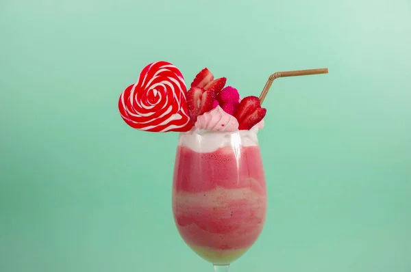 Nahaufnahme eines köstlichen hausgemachten extremen Milchshakes aus Erdbeeren mit Milchschaum mit einem Plastikstroh und einer rosafarbenen Brombeerbonbon mit einem Herzbonbon darüber, vor einem weichen blauen Hintergrund — Stockfoto