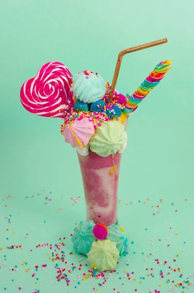 Délicieux milkshake extrême de fraise avec des dragees et un bonbon à la mûre sur une mousse de lait avec une paille en plastique et un bonbon au cœur sur le dessus, avec quelques dragees éparpillés à la base, dans un bleu doux — Photo