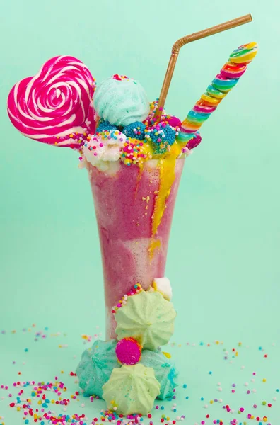 Délicieux milkshake extrême de fraise avec des dragees et un bonbon à la mûre sur une mousse de lait avec une paille en plastique et un bonbon au cœur sur le dessus, avec quelques dragees éparpillés à la base, dans un bleu doux — Photo
