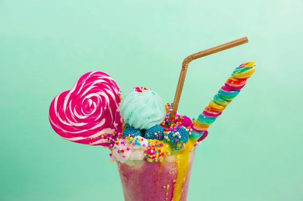 Dekat milkshake ekstrim lezat stroberi dengan dragees dan permen blackberry di atas busa susu dengan sedotan plastik dan permen jantung di atas, di latar belakang biru lembut — Stok Foto