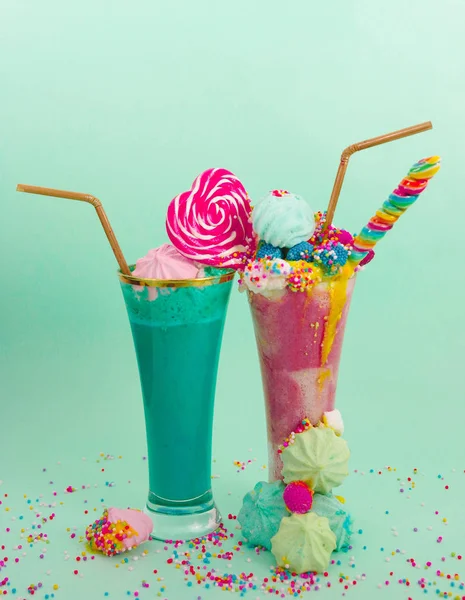 Dua rakus buatan sendiri milkshake ekstrim, dengan permen blackberry di atas busa susu dan permen pelangi di atas dengan sedotan plastik di latar belakang biru lembut — Stok Foto