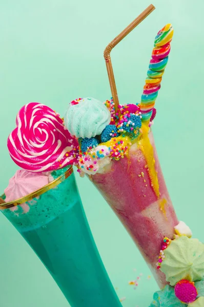 Deux elicious milkshake maison extrême, avec un bonbon à la mûre sur une mousse de lait et un bonbon arc-en-ciel sur le dessus avec une paille en plastique dans un fond bleu doux — Photo