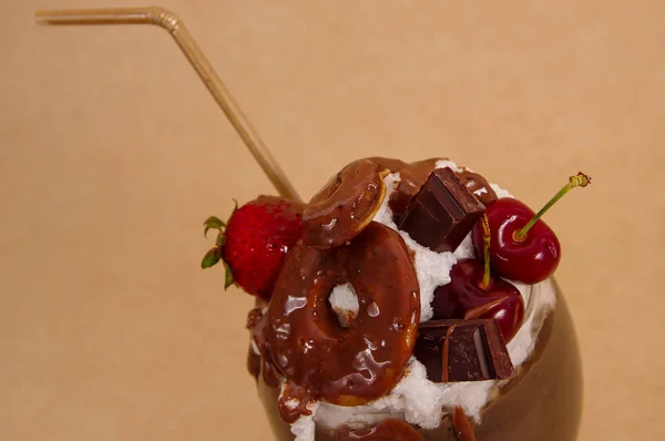Närbild på choklad överseende extrema milkshake med brownie kaka, jordgubbar, körsbär och en plast halm med mjölkskum på toppen i en mjuk brun bakgrund — Stockfoto