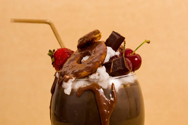 Närbild på choklad överseende extrema milkshake med brownie kaka, jordgubbar, körsbär och en plast halm med mjölkskum på toppen i en mjuk brun bakgrund — Stockfoto