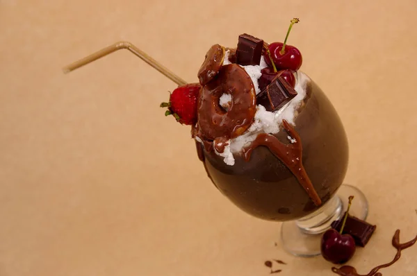 Chocolate indulgent extreme milkshake with brownie cake, strawberries, cherries, and a plastic straw with milk foam on top in a soft brown background — Stock Photo, Image