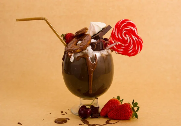 Chocolate indulgent extreme milkshake with brownie cake, strawberries, cherries, candy with heart form, a plastic straw with milk foam on top in a soft brown background — Stock Photo, Image