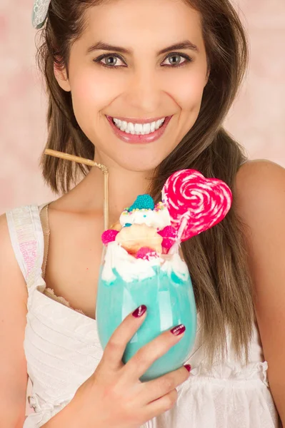 Sorridente giovane bella donna con un gustoso frullato di latte blu in studio di moda, con una caramella cuore, paglia di plastica su una schiuma di latte in cima, in uno sfondo rosa — Foto Stock