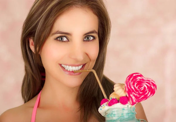 Sorridente giovane bella donna, indossa una valigia rosa, tenendo un gustoso frullato di latte blu in studio di moda, con una caramella cuore, paglia di plastica su una schiuma di latte in cima, in uno sfondo rosa — Foto Stock