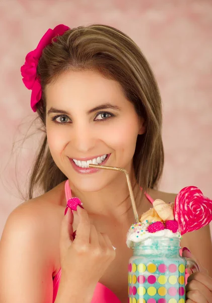 Primo piano di una giovane bella donna sorridente, che indossa una valigia rosa, con in mano un gustoso frullato di latte blu in stile studio, con una caramella al cuore, paglia di plastica su una schiuma di latte in cima, su uno sfondo rosa — Foto Stock