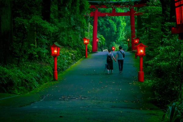 Pareja caminando y sosteniendo sus manos en vista nocturna del acercamiento al santuario de Hakone en un bosque de cedro. Con muchas linternas rojas iluminadas y una gran puerta torii roja —  Fotos de Stock