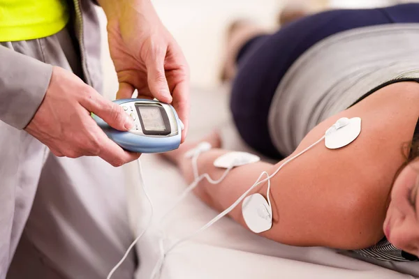 Närbild av en personlig tränare håller med å ena sidan den elektriska maskinen och sätta electrostimulator elektroderna i armen av en kvinnlig deportist, upp och ned på en säng — Stockfoto