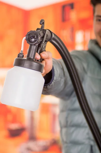 Verschwommen gutaussehender lächelnder Handwerker mit der Malerpistole in der Hand, ein in einer grauen Jacke vor verschwommenem Hintergrund — Stockfoto