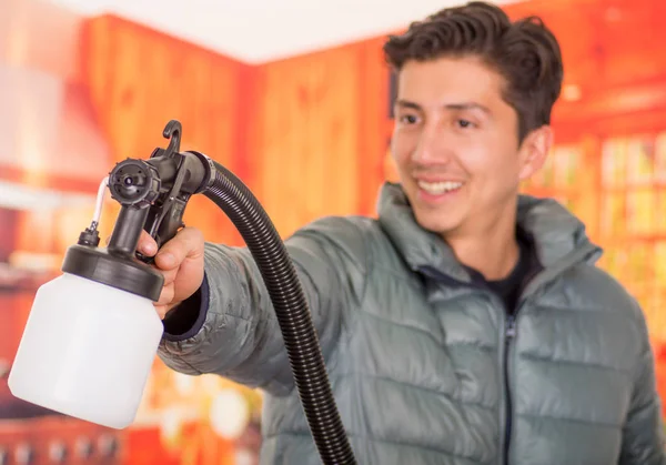 Blurred handsome smiling handyman holding in his hands the painting spray gun, an wearing a gray jacket in a blurred background