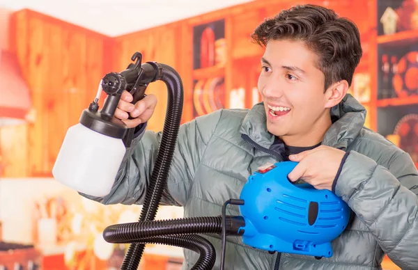 Gros plan d'un bel homme souriant tenant dans ses mains le pistolet à peinture, portant une veste grise dans un fond flou — Photo