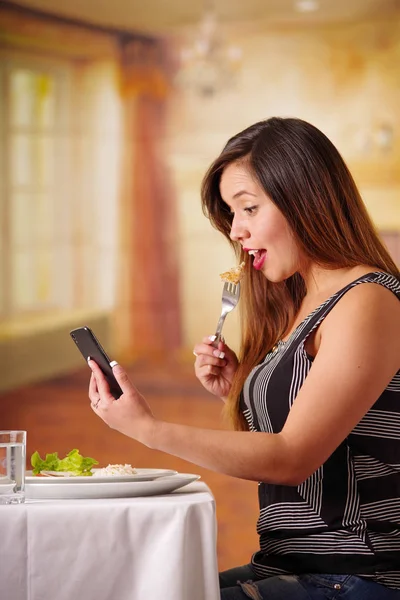 Retrato de una hermosa mujer sorprendida comiendo sola en un restaurante y revisando su teléfono celular, en un fondo borroso, vista de perfil — Foto de Stock