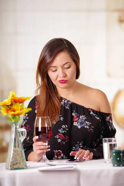 Porträt einer schönen eleganten Frau mit langen Haaren, die allein einen Rotwein trinkt und in einem Restaurant vor verschwommenem Hintergrund ihr Handy in der anderen Hand hält — Stockfoto