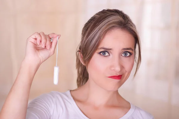 Young beautiful woman with a bored face, holding a menstruation cotton tampon in her hand, in a blurred background — Stock Photo, Image