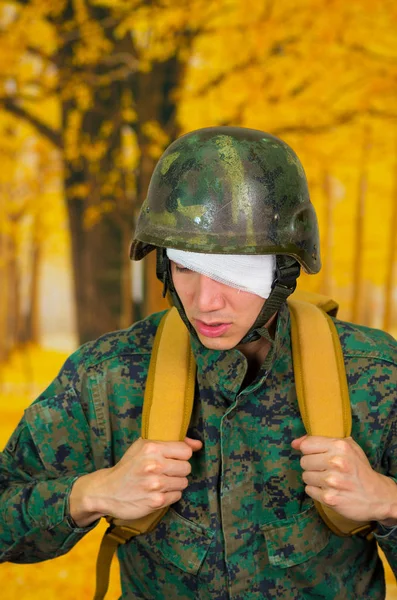 Bonito jovem soldado vestindo uniforme sofrendo de estresse, com uma bandagem branca em torno de sua cabeça e cobrindo seu olho, com uma mochila em um fundo amarelo enterrado — Fotografia de Stock