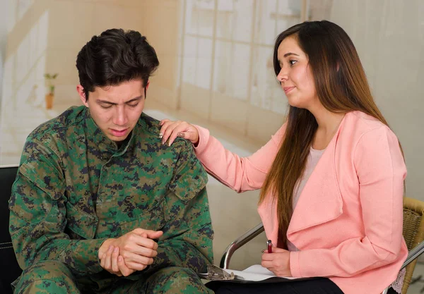 Красивый молодой солдат в форме, страдающий от стресса, с женщиной-терапевтом, оказывающей поддержку пациенту в медицинском центре здоровья, вспоминая ужасы войны, на зарытом фоне — стоковое фото
