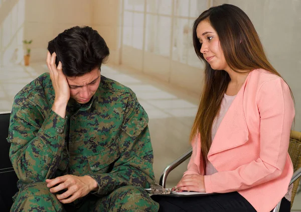 Handsome young soldier wearing uniform suffering from stress, with a woman therapist in a medical health center, touching his head with one hand and Remembering the horrors of war, in a burred — Stock Photo, Image